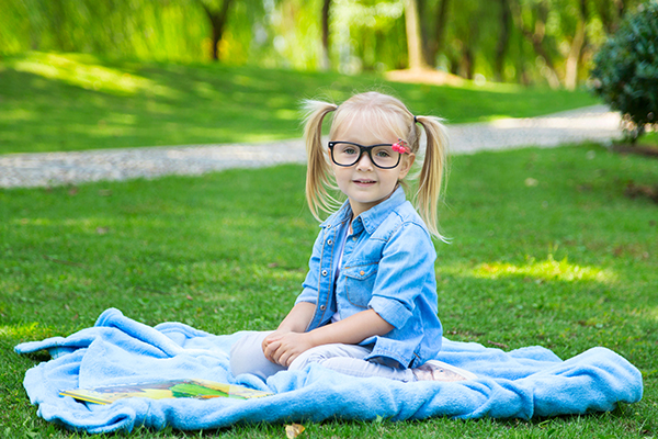 Kilkuletnia dziewczynka w okularach siedzi na kocu rozłożonym na trawie. Uśmiecha się. 
