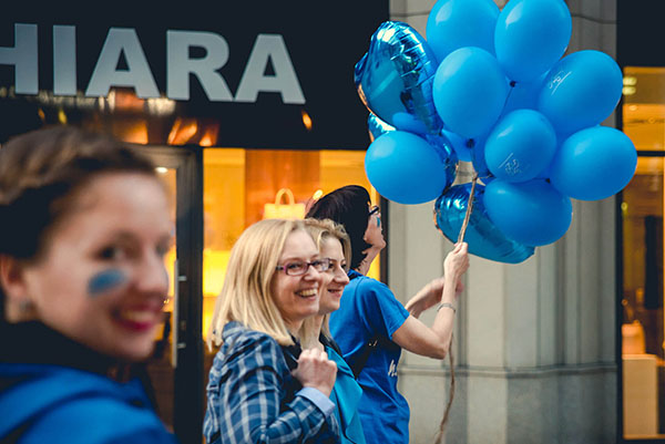Grupa roześmianych osób ubranych na niebiesko. Jedna trzyma w rękach dużo niebieskich balonów.