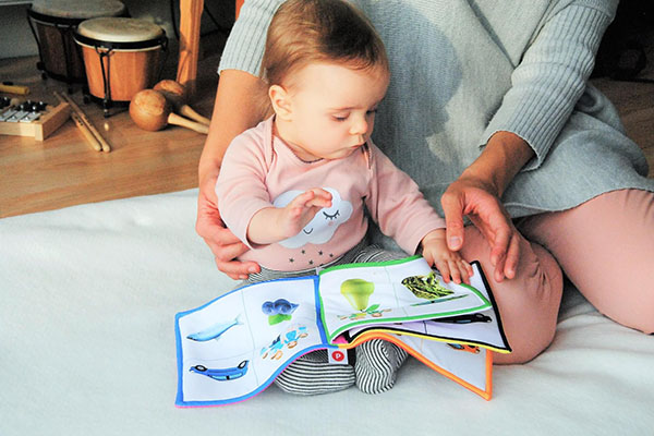 Małe dziecko przegląda kolorową książeczkę. Siedząca obok kobieta otacza je ramieniem. 